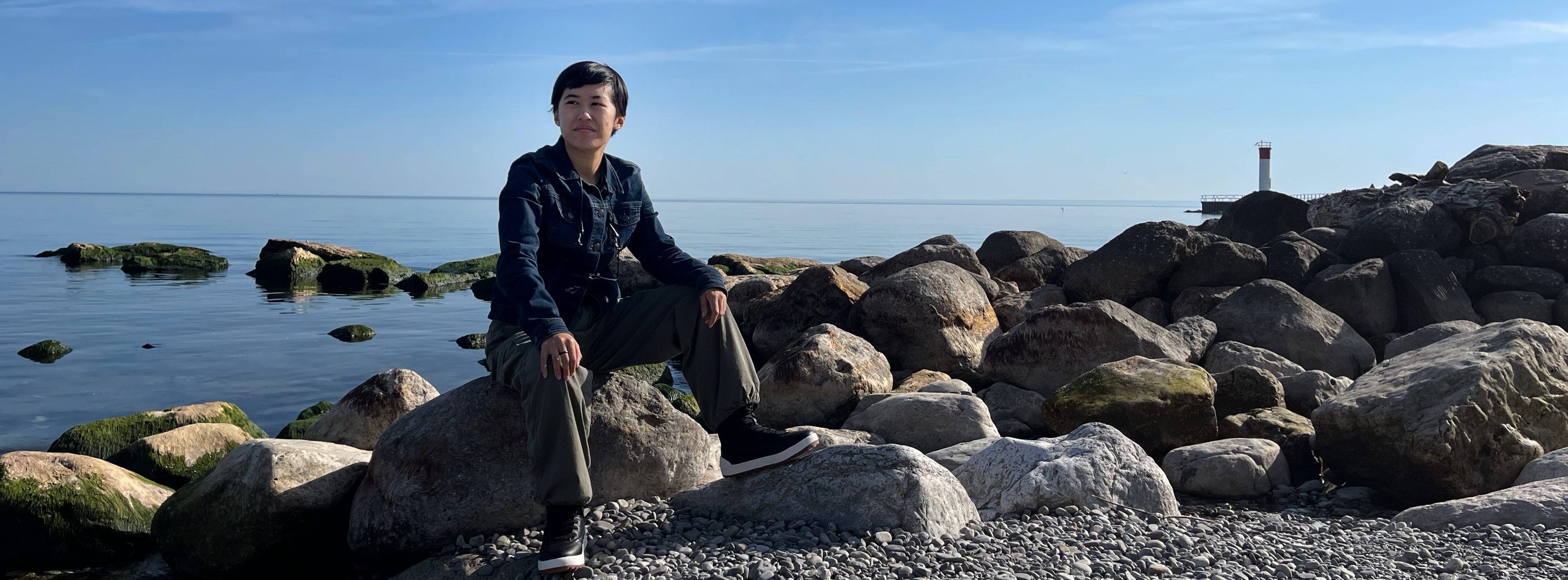 A photo of M. Li Zeng sitting on a rock next to a lake. M. is a genderqueer Chinese person with short black hair. In the photo, they are wearing a jean jacket, green cargo pants, and black boots. They look off to one side with a neutral expression.